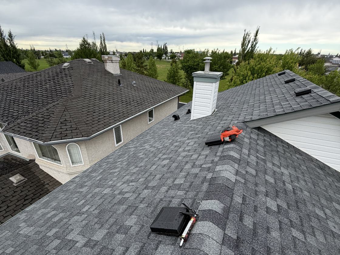A SureSky re-roof underway: fresh asphalt shingles laid across an Alberta rooftop, with a roofing nailer and caulking gun on the deck beside the chimney