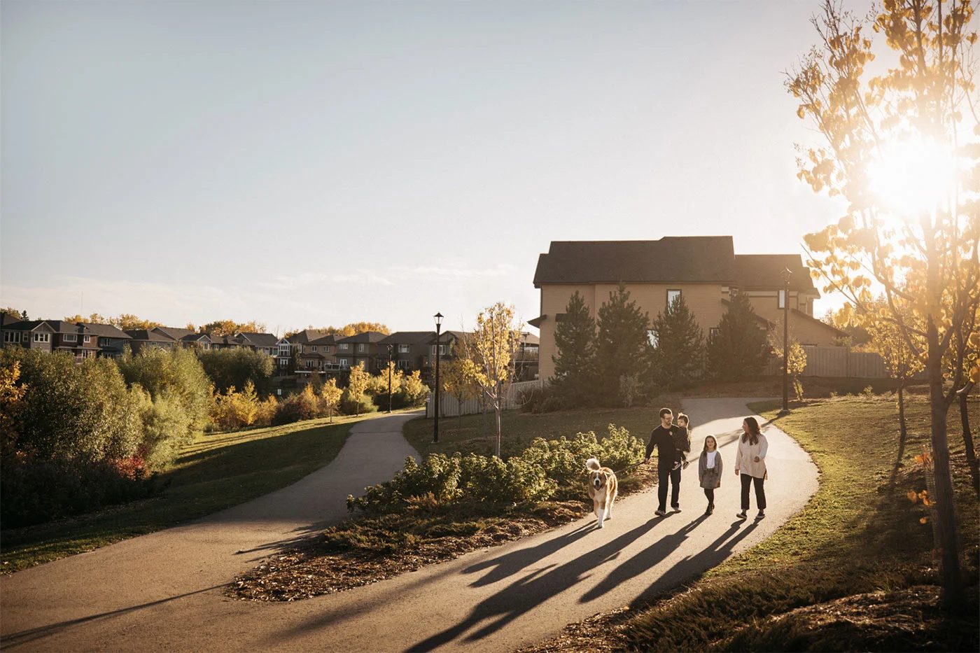 A family walking their dog on a neighbourhood path at golden hour, homes in the background