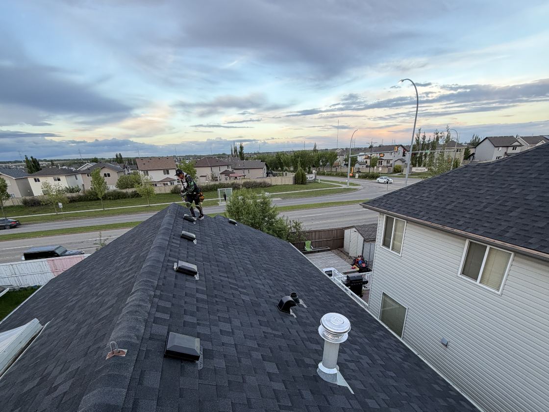 A SureSky roofer standing on the ridge of an asphalt shingle roof, checking the run of vents above an Alberta neighbourhood at dusk